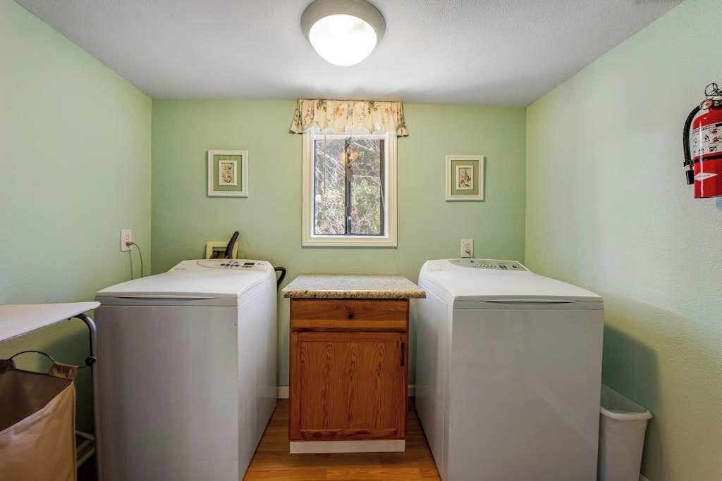 A laundry room with two washing machines, a wooden cabinet, a window with a floral valance, and a fire extinguisher on the wall.