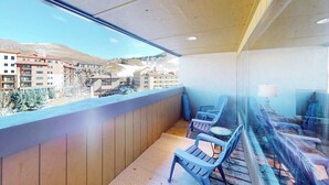 Un balcón con vistas a edificios y montañas, suelo de madera y sillas azules.