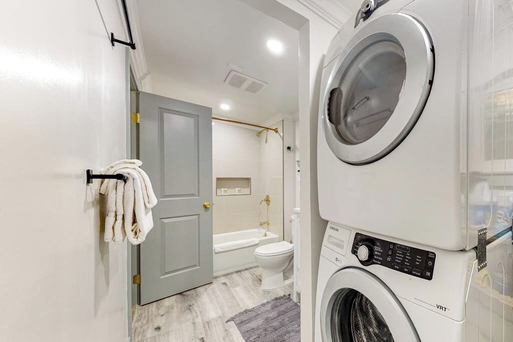 Une buanderie équipée d’un lave-linge et d’un sèche-linge superposés, une porte donnant sur une salle de bain avec une baignoire et des toilettes, et un porte-serviettes avec des serviettes.