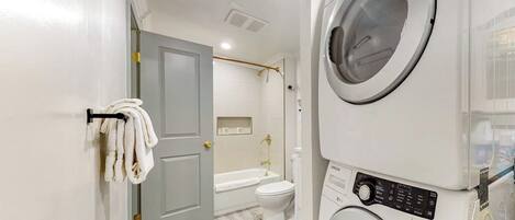 Une buanderie équipée d’un lave-linge et d’un sèche-linge superposés, une porte donnant sur une salle de bain avec une baignoire et des toilettes, et un porte-serviettes avec des serviettes.