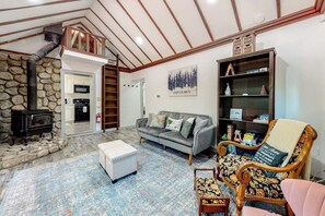 A cozy living room with a stone fireplace, a sofa, a patterned armchair, and a bookshelf. - Forest-view cabin with deck, grill, wood stove, washer/dryer, & fast WiFi (Crestline)