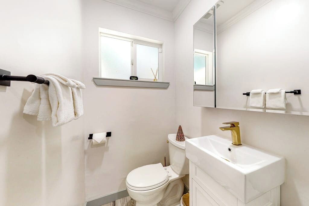 A bathroom with a white toilet, a gold faucet sink, a towel rack, and a window above the toilet.