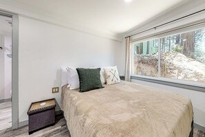 A bedroom with a bed, a window with a view of a forest, and a small bedside table with a lamp. - Forest-view cabin with deck, grill, wood stove, washer/dryer, & fast WiFi (Crestline)