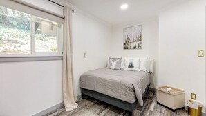 Een slaapkamer met een bed, een raam met gordijnen, een foto aan de muur en een prullenbak.
