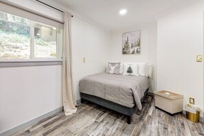 A bedroom with a bed, a window with curtains, a picture on the wall, and a trash bin. - Forest-view cabin with deck, grill, wood stove, washer/dryer, & fast WiFi (Crestline)