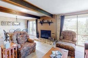 A cozy living room with a fireplace, patterned furniture, and a view of trees through large windows. - Beautiful mountain home steps to ski slopes with fireplace (Durango)