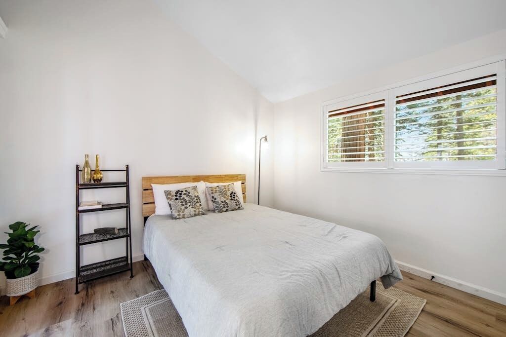 A bedroom with a bed, a bedside shelf, a window with blinds, and a plant.