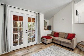 Ein Zimmer mit einer Schiebetür, einem beigen Sofa mit roten Kissen, einem kleinen Hocker und einem weißen Teppich auf Holzdielen.
