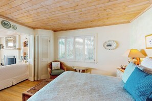 A bedroom with a wooden ceiling, a bed with a patterned bedspread, a bedside table with a lamp, a chair, a small table, and a window with shutters.