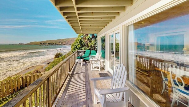 Une terrasse en bois avec des chaises blanches, une balustrade en verre et une vue sur la plage et l’océan.