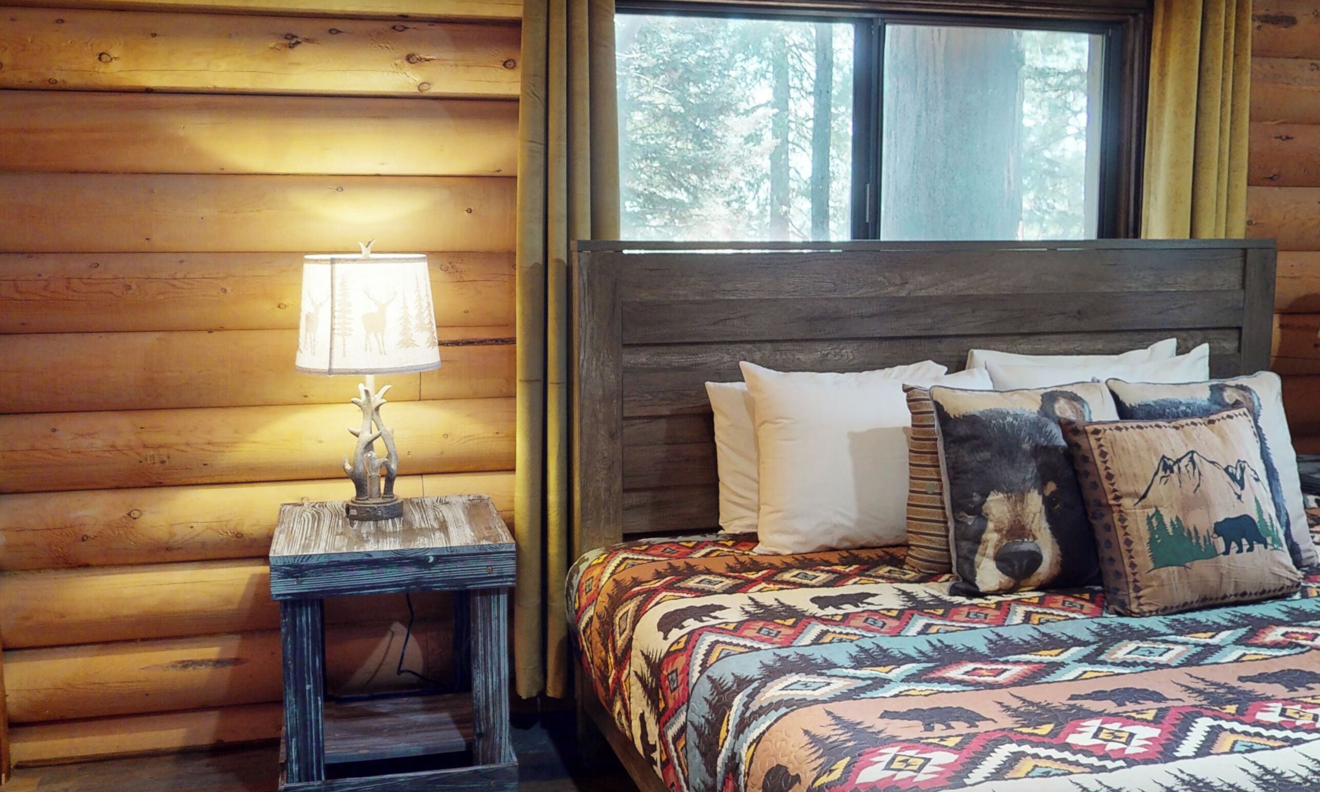 A wooden cabin room with a bed, a nightstand with a lamp, and a window with a view of trees.