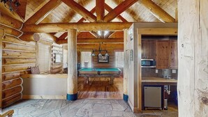A log cabin interior with a stone floor, wooden beams, a billiards table, and a kitchen area.