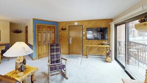 Ein Zimmer mit einer Holzwand, einem Karo-Sessel, einem Fernseher und einem großen Fenster.