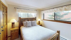 Ein Schlafzimmer mit einem hölzernen Kopfende, zwei Nachttischen mit Lampen, einem großen Fenster mit Vorhängen und Blick auf Bäume.