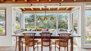 A dining area with a wooden table and chairs, a sign indicating Pebble Beach 5 miles away, and large windows offering a view of the outdoors. - Surf-side monthly rental with ocean views, beach access & private hot tub (Carmel-by-the-Sea)