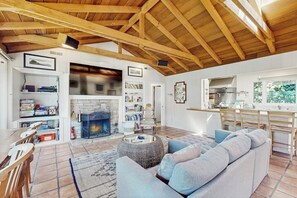 A spacious living area with a fireplace, a sofa, a dining table, and a kitchen in the background. - Surf-side monthly rental with ocean views, beach access & private hot tub (Carmel-by-the-Sea)