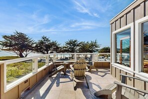 A balcony with outdoor seating, a view of the sea, and a building with large windows. - Surf-side monthly rental with ocean views, beach access & private hot tub (Carmel-by-the-Sea)