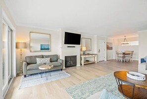 A modern living room with a sofa, coffee table, fireplace, TV, dining area, and a large window. - Ocean-view condo in Santa Barbara steps to beach with pool & balcony (Santa Barbara)