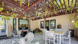 Un patio al aire libre con mobiliario de mimbre blanco, una pérgola con enredaderas y una zona de barbacoa.