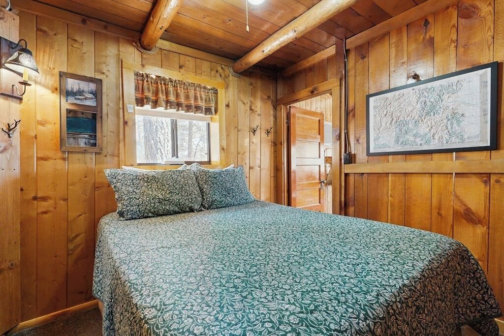 A wooden cabin room with a bed, a window with curtains, a framed map, and a wall-mounted lamp.