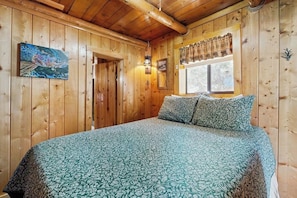 A wooden cabin interior with a bed, a door, a window with curtains, and a wall-mounted lamp.