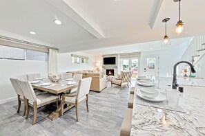 Eine moderne Küche mit Kochinsel, ein Essbereich mit Holztisch und -stühlen sowie ein Wohnbereich mit Sofa und Fernseher.