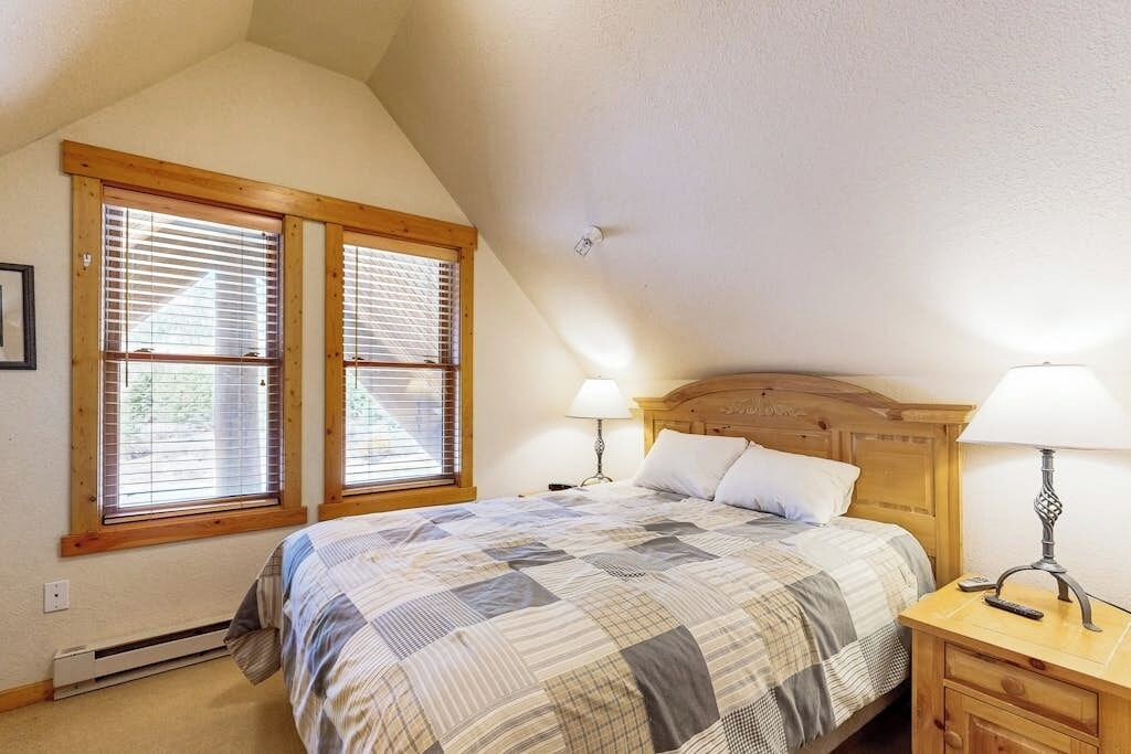 Une chambre avec un plafond en pente, une tête de lit en bois, une couvre-lit matelassée, deux lampes de chevet et une fenêtre avec des stores.
