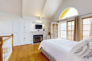 A bedroom with a fireplace, a large bed, a TV mounted above it, and a window with curtains.