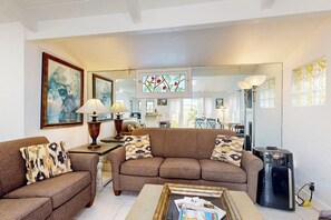 A living room with a sofa, two side tables with lamps, a coffee table with magazines, a mirror, and a refrigerator.