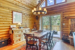 Ein Holzhütten-Interieur mit Esstisch, Stühlen, einer Deckenleuchte, einem Kamin und einem großen Fenster.