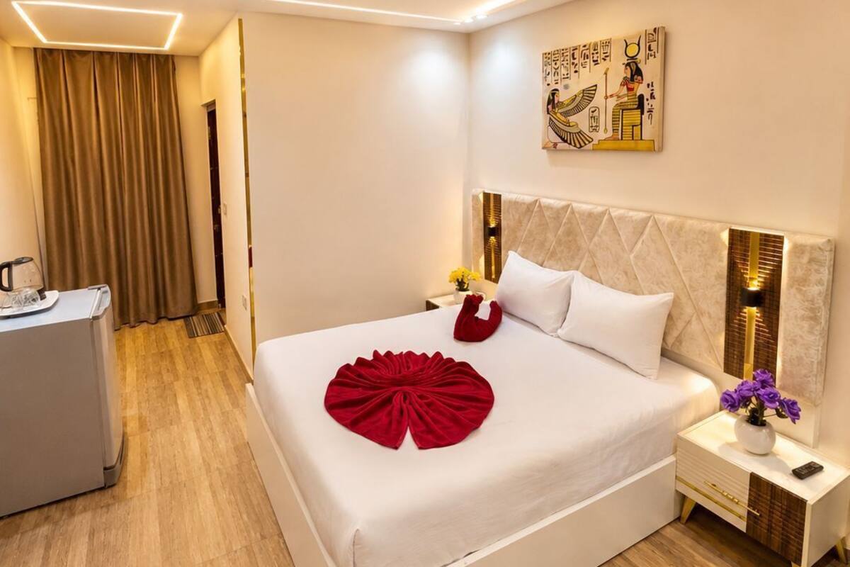 A hotel room with a bed, bedside table, nightstand, and a painting on the wall.