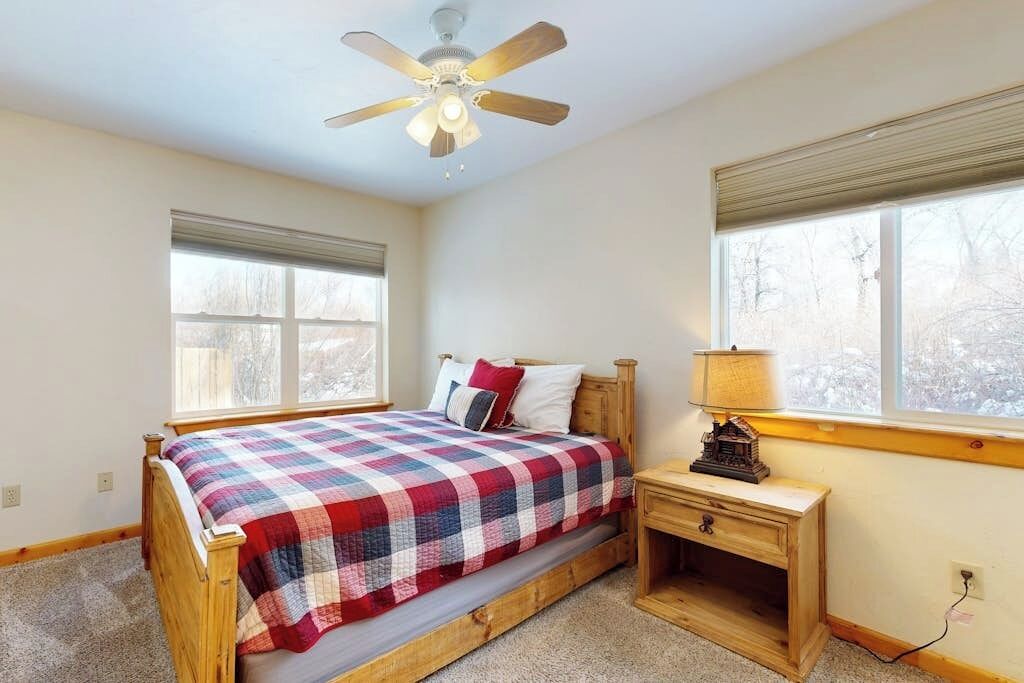 Ein Schlafzimmer mit einem hölzernen Bett, einem Deckenventilator, einem Nachttisch mit Lampe und einem Fenster mit Blick auf Bäume.