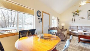 A living room with a wooden dining table, sofas, and a large window with a view of trees.
