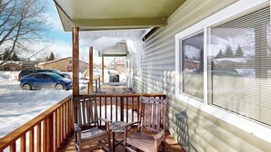 A covered porch with wooden chairs and a table, a window with blinds, and a snowy driveway.