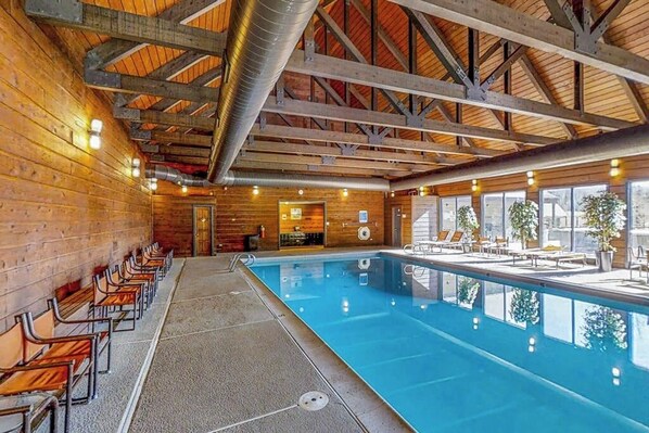 An indoor swimming pool with wooden walls and ceiling, benches along the poolside, and a view of the outside.