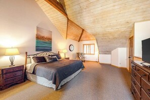 Ein Schlafzimmer mit geneigter Decke, einem Bett mit dunkel farbenfroher Bettdecke, einem Nachttisch mit Lampe, einem Sessel, einer Kommode und einem Fernseher.