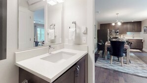 Une salle de bain moderne avec un grand miroir, un lavabo blanc et une chaise aux tons foncés dans l’espace repas adjacent.