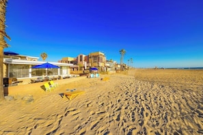 A beachside hotel with outdoor seating, palm trees, and a clear blue sky. - Oceanfront Beach Cottage On the Sand + Unobstructed Views (Newport Beach)