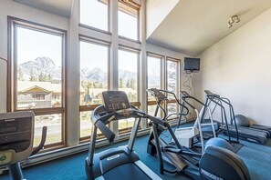 Ein Fitnessstudio mit Laufbändern, einem Ergometer und einem Rudergerät, großen Fenstern mit Bergblick und einem an der Wand montierten Fernseher.