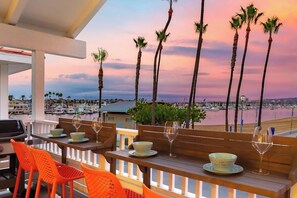 Eine Terrasse mit Holztischen und orangefarbenen Stühlen mit Blick auf einen Hafen voller Boote und Palmen vor einem Sonnenuntergangshimmel.