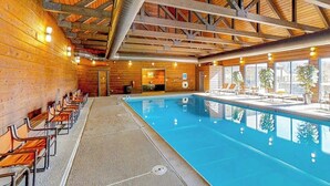 Une piscine intérieure aux murs et au plafond en bois, avec des bancs disposés le long du bord de la piscine et une vue sur l’extérieur grâce à de grandes fenêtres.