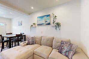 A living room with a beige sectional sofa, a dining table with chairs, and a painting of a sailboat on the wall. - Cozy, Single-Level Getaway with Fast WiFi, Kitchenette, & A/C - Dogs Welcome (Newport Beach)