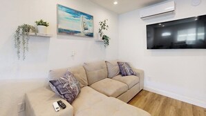 A living room with a sofa, a painting, a mounted TV, and an air conditioning unit. - Cozy, Single-Level Getaway with Fast WiFi, Kitchenette, & A/C - Dogs Welcome (Newport Beach)