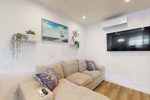 A living room with a sofa, a painting, a mounted TV, and an air conditioning unit. - Cozy, Single-Level Getaway with Fast WiFi, Kitchenette, & A/C - Dogs Welcome (Newport Beach)