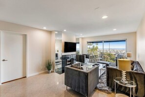 A modern living room with a fireplace, a flat-screen TV, a sofa, and a large window offering a city view. - Ocean-view condo, balcony, clubhouse, gym, pool, hot tub, snowbird haven (Newport Beach)