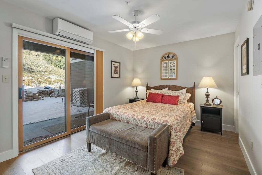 Quarto com cama, duas mesas de cabeceira com luminárias, um ventilador de teto, um banco e uma porta de vidro deslizante para área externa.
