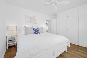 Une chambre à coucher blanche, avec un lit, une table de chevet, une lampe, un ventilateur de plafond et une armoire.