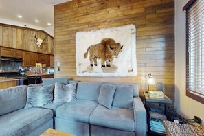 Ein Wohnzimmer mit grauem Sofa, einer Holzwand mit angebrachtem Tierkopf und einer Küche im Hintergrund.