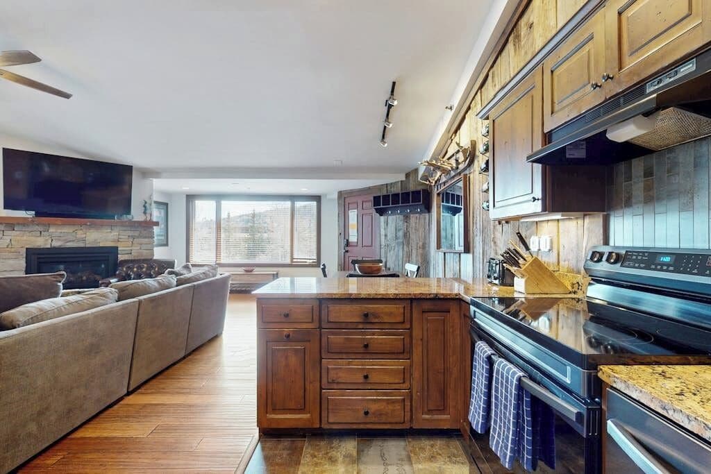 A modern kitchen with wooden cabinets, a built-in island, a large stove, and a flat-screen TV mounted above the fireplace.