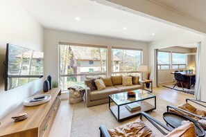 Ein großzügiges Wohnzimmer mit einer Couch, einem Couchtisch und Ledersesseln. Links befindet sich ein Fernseher und auf der anderen Seite ein Essbereich mit Tisch und Stühlen.